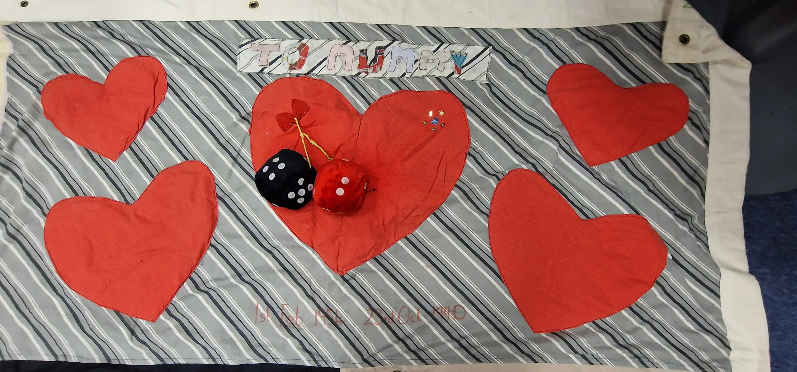 An image showing a striped quilt with red hearts in it. Written at the top is 'To Mummy', and at the bottom are the dates 1st February 1954 - 23rd October 1991.